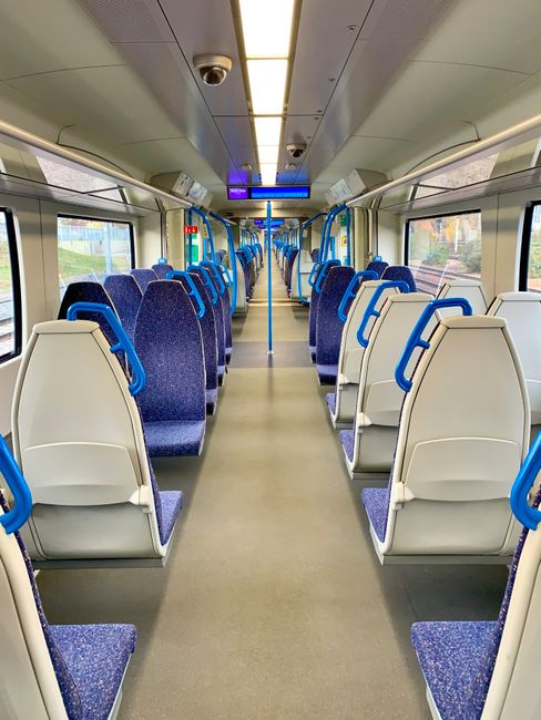Interior view of a train carriage with rows of blue upholstered seats featuring a swirling pattern, arranged in pairs along the aisle. The floor is a smooth, light gray surface, appearing clean and we