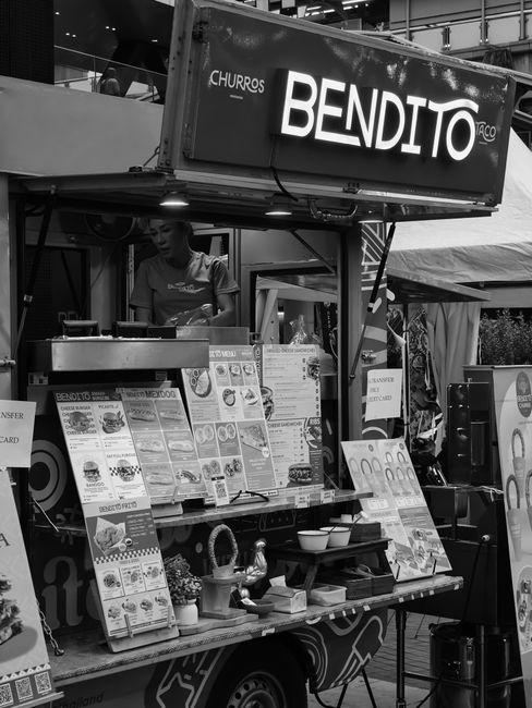 A male food vendor in a red polo shirt with the Spitalfields Market logo is preparing food at a stall. The stall features a stainless steel surface with a row of small, round food items, likely snacks